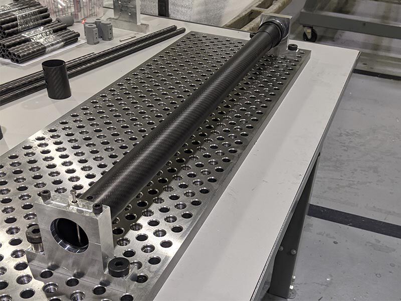 Image shows work surface with metallic tooling. A carbon fiber tube is held in place for precision drilling and assembly.